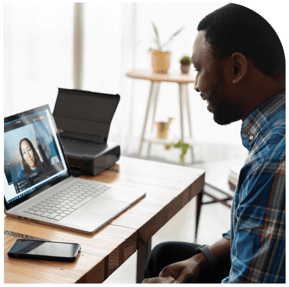 A man sits at a wooden desk, smiling at a laptop during a video call. A mobile phone and printer are on the desk, and a small table with potted plants is in the background.