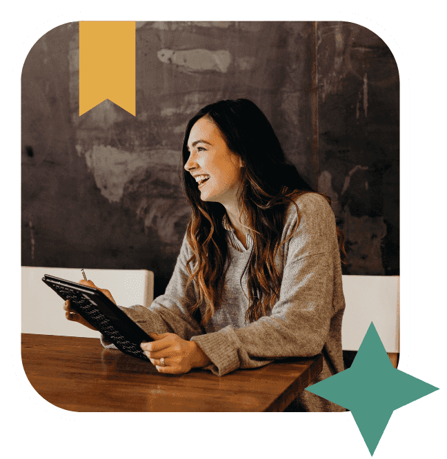 A woman with long brown hair, wearing a grey jumper, sits at a wooden table smiling and holding a tablet with a stylus in a cosy, modern setting. Decorative shapes frame the photo.