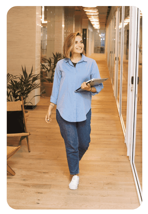A person wearing a light blue shirt, jeans, and white trainers walks down a corridor with wooden floors, holding a laptop and smiling. Potted plants and glass office walls are visible in the background.