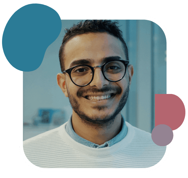 Young man with glasses and a friendly smile, wearing a white sweater. Background is softly blurred, featuring blue and pink abstract shapes.