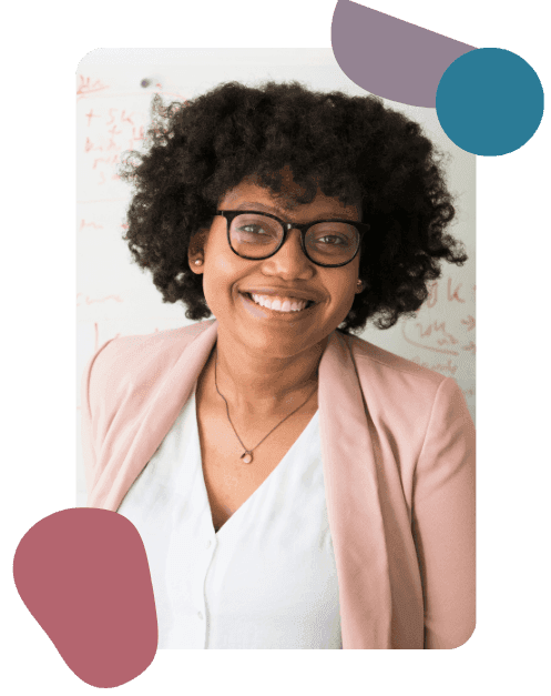 Smiling woman with curly hair and glasses, wearing a light pink blazer and white top, stands against a whiteboard backdrop. The mood is friendly and professional.