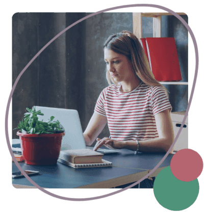 A woman in a striped shirt works on a laptop at a desk with a notebook and a potted plant.