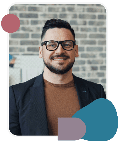 Man with short dark hair, beard, and glasses wearing a brown shirt and black blazer stands smiling in front of a brick wall, with colored graphic shapes overlayed.