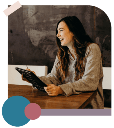 Woman with long brown hair, wearing a grey sweater, sits at a wooden table holding a tablet and smiling. Dark chalkboard wall in the background, abstract shapes decorate the image corners.