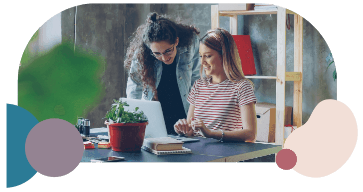 Two women smiling and collaborating at a desk with a laptop, surrounded by notebooks and a potted plant, conveying a positive, creative atmosphere.