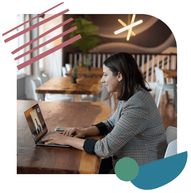 Woman in checkered blazer sits at a wooden table, engaged in a video call on a laptop. The room has modern decor and a warm ambiance.