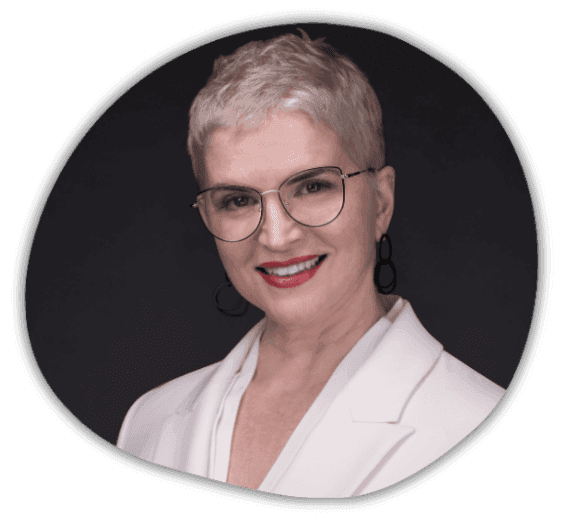 A person with short, light hair wearing glasses, a white blazer, and hoop earrings smiling against a dark background.