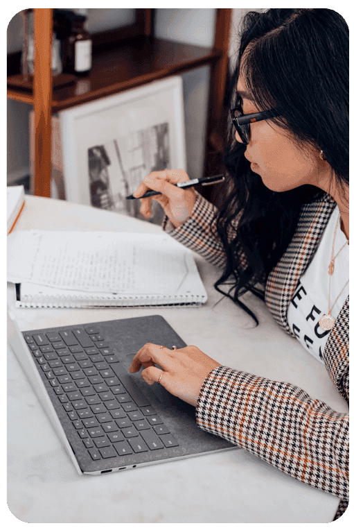 Seen over her shoulder, a woman in a checked blazer works at a laptop with a pen in her hand and an open ring binder beside her.