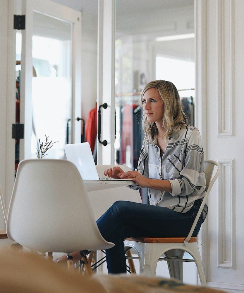 A woman in a striped shirt sits at a small white table in a bright room, working at a laptop.