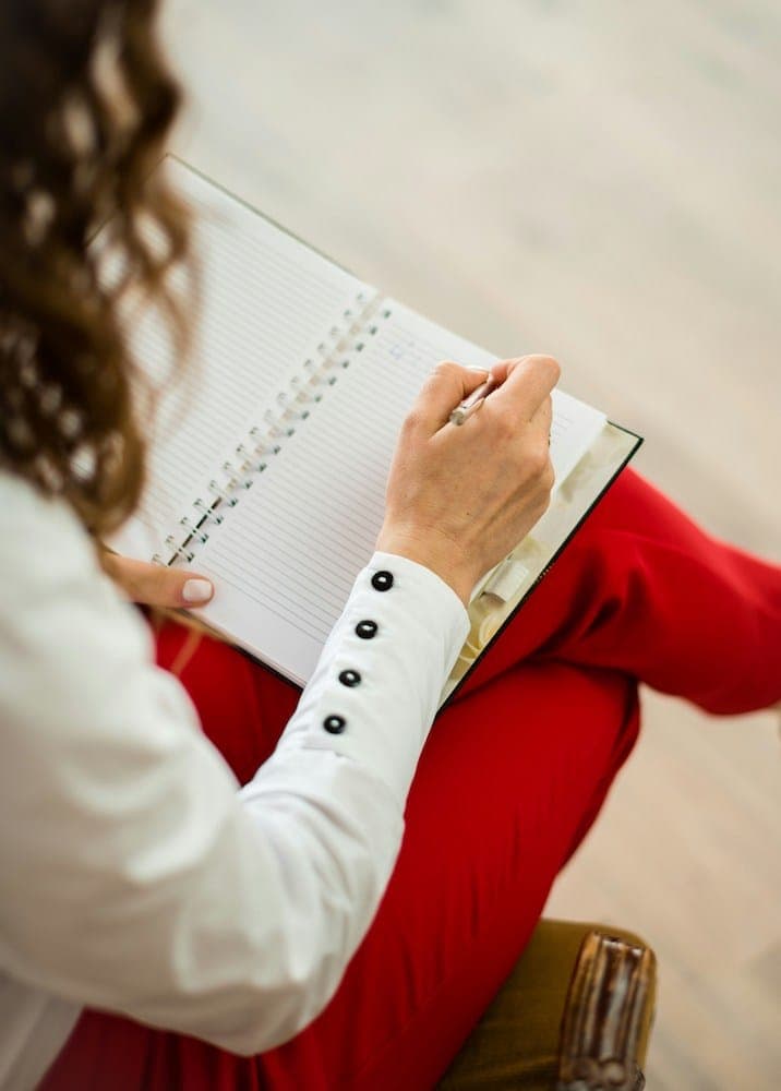 Seen from above, a woman in a red dress and white shirt writes in a spiral notebook resting on her lap.
