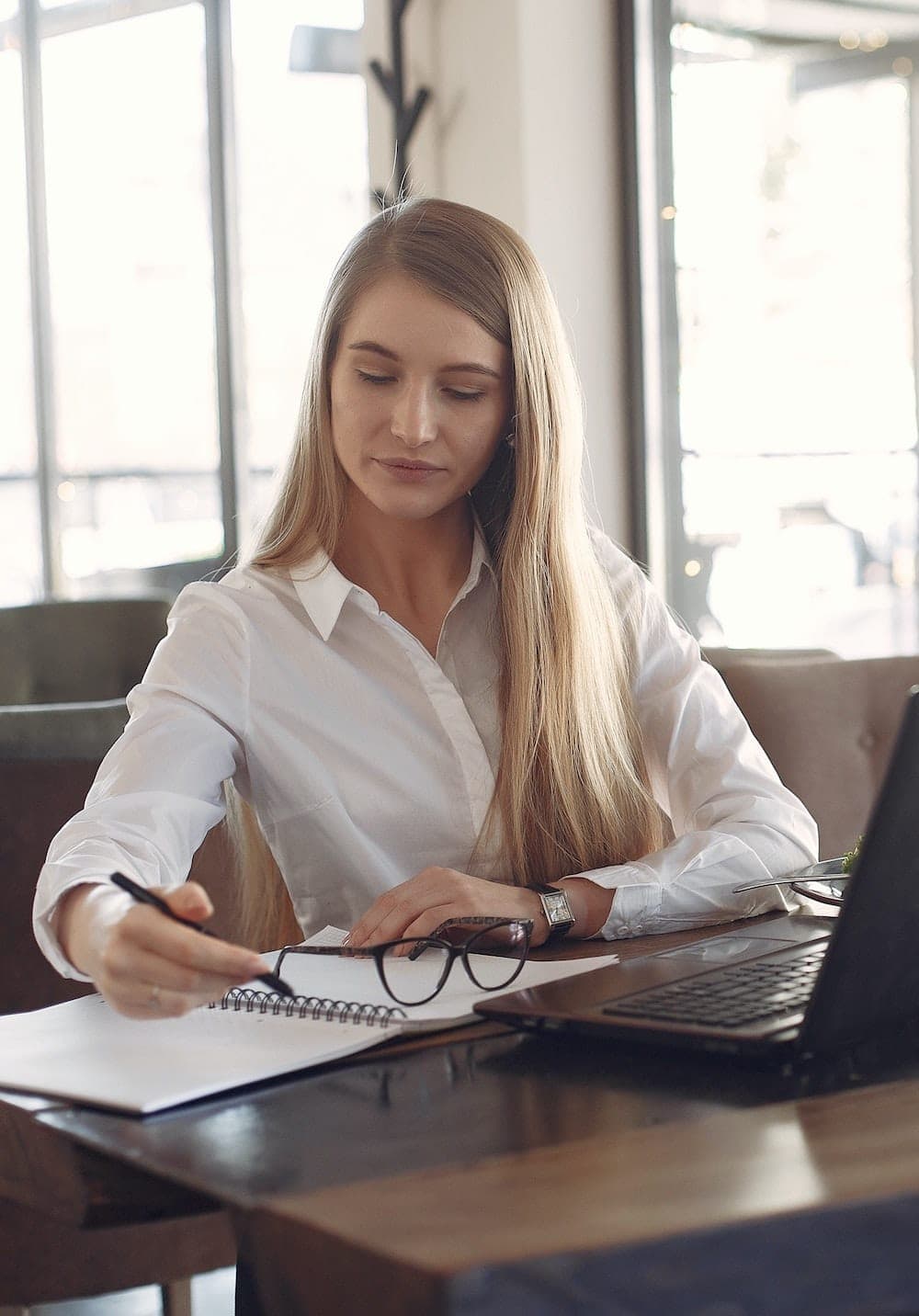 A woman in a white shirt writes in a spiral notebook at a cafe table, with a laptop and her glasses beside her.