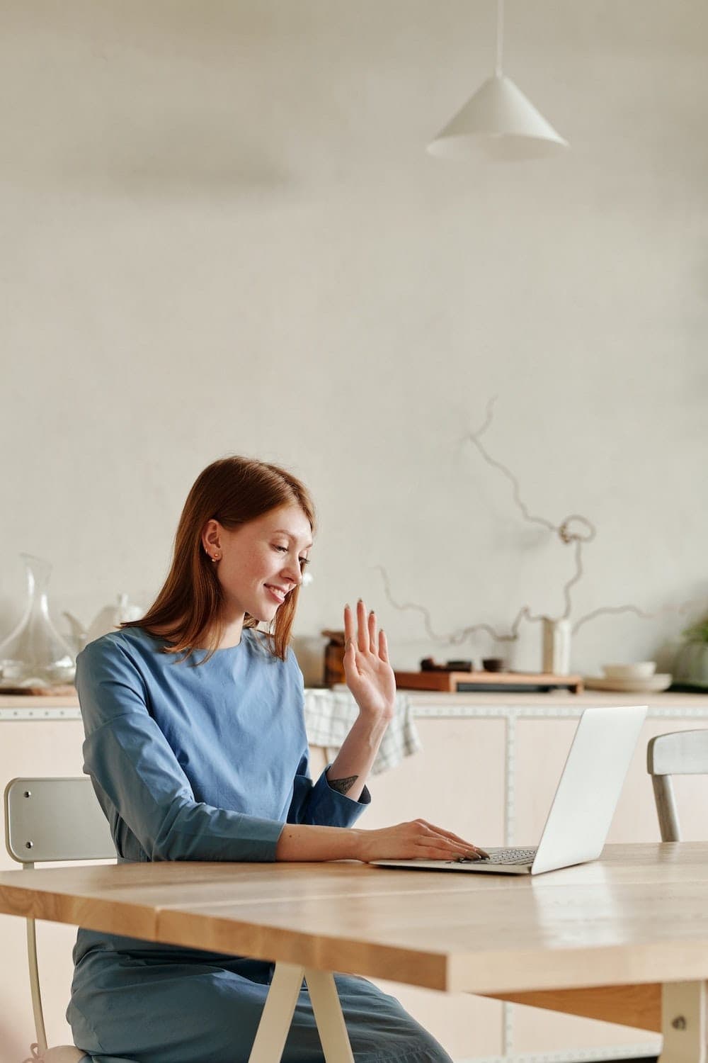 A woman in a blue top sits at a wooden kitchen table, waving at her open laptop during a video call.