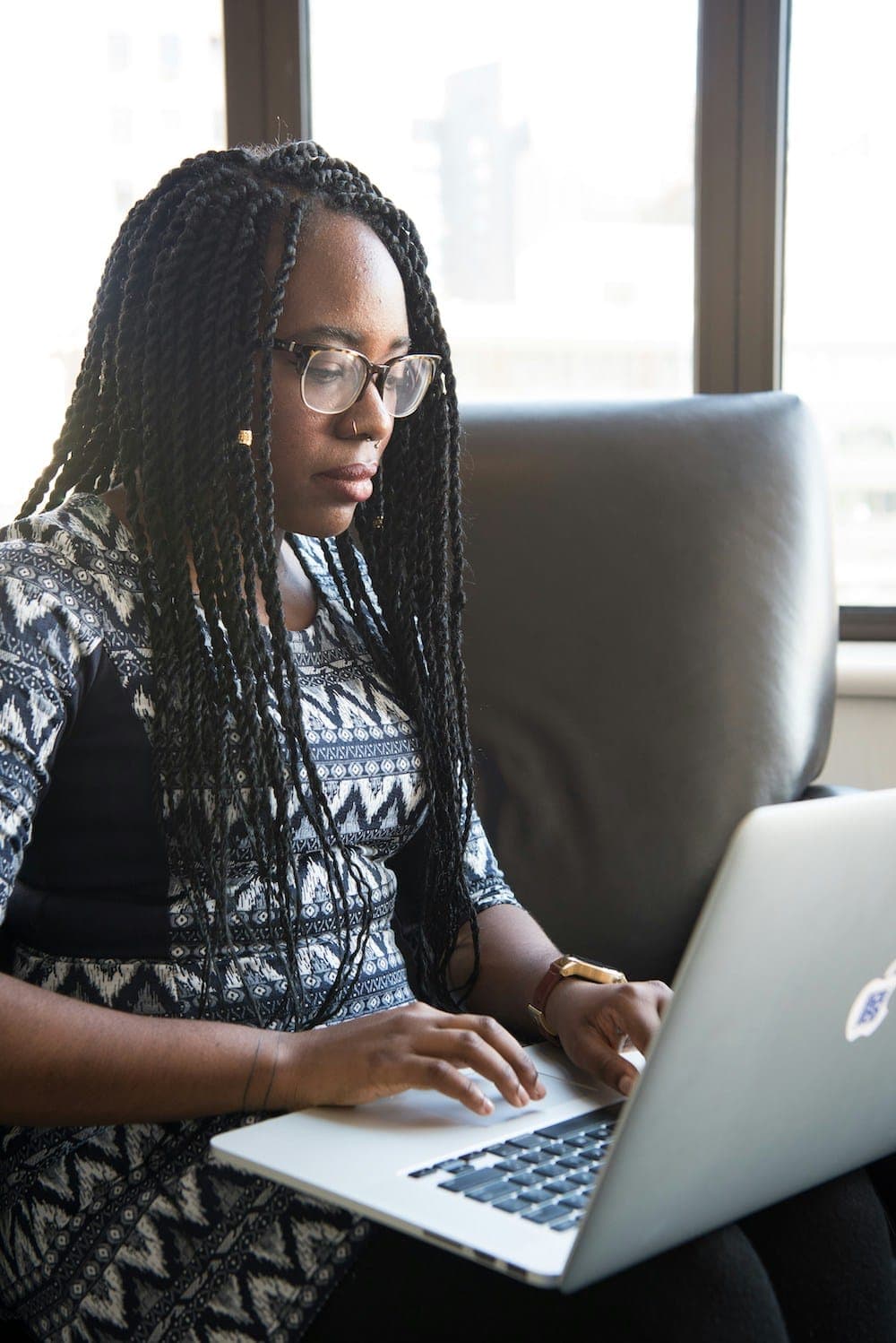 A woman with long braids and glasses works at a laptop on her lap, beside a window overlooking a city.