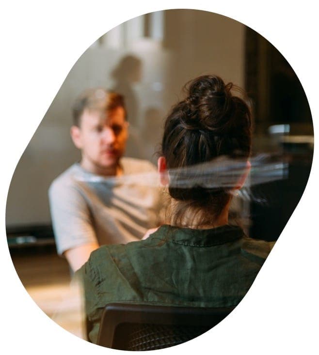 Seen past the shoulder of a woman with her hair in a bun, a bearded man in a pale t-shirt leans forward to listen to her across a table.