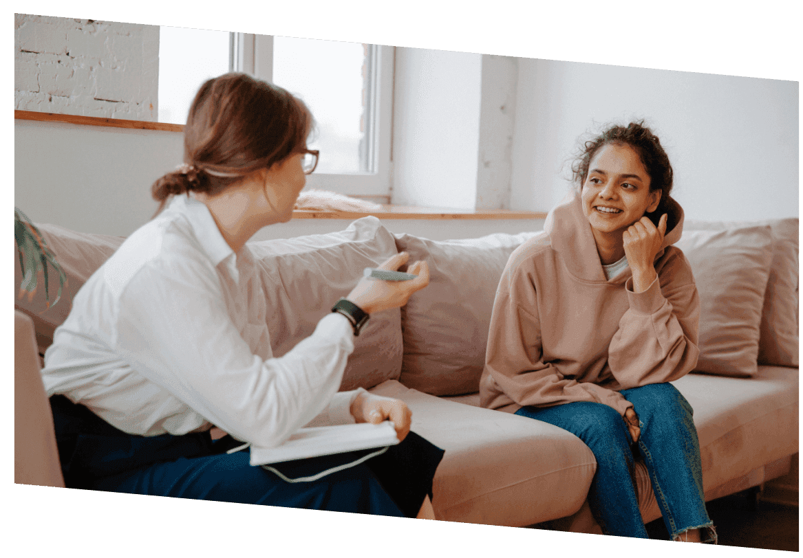 Two women talk on a pink sofa in a bright room; one in a white shirt holds a pen over an open notebook, the other rests her chin on her hand and smiles.
